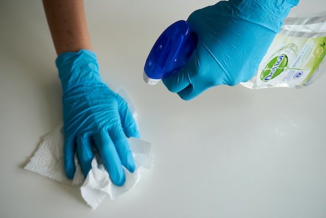 hands in blue rubber gloves using spray bottle and paper towel to clean white surface